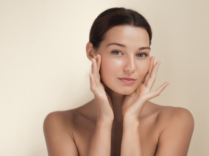 A woman with radiant skin looks into the camera, holding her hands on either side of her face.