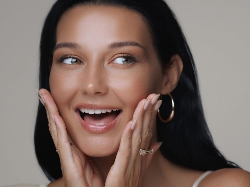 A woman with glowing skin smiles and looks to the side, with her hands on either side of her face in a show of surprise.