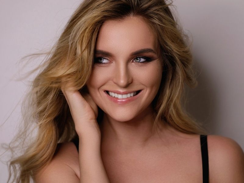 A blonde woman smiles into the camera, with one hand in her hair.