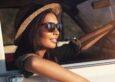 A woman in a summer hat and sunglasses peers out of a car.