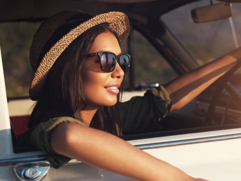 A woman in a summer hat and sunglasses peers out of a car.