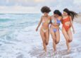 Three women walk on the beach in their swimsuits.